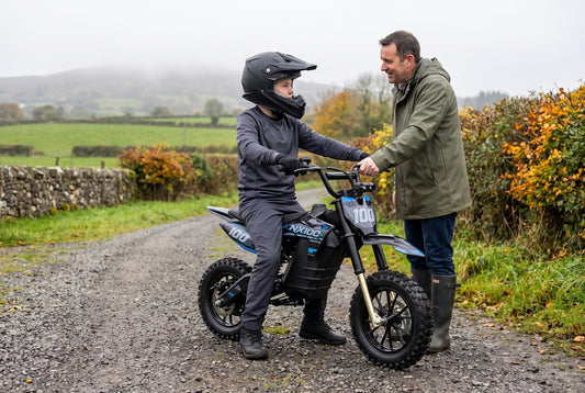 Parent beside a young rider in a full-face helmet sitting on a Novi NX100 electric dirt bike on a farm lane in County Tyrone