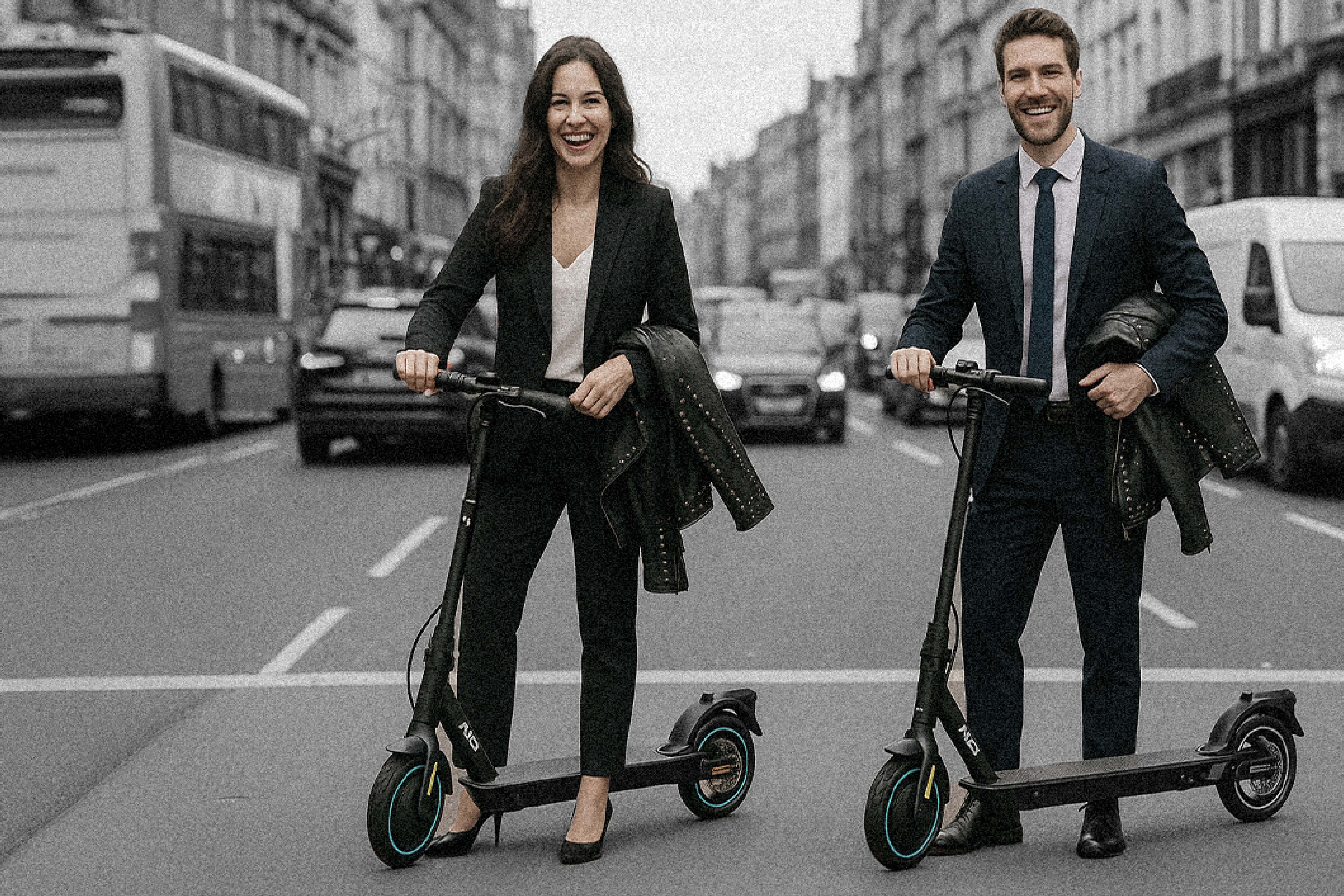 Two people riding electric scooters on a city street with buildings and vehicles in the background.